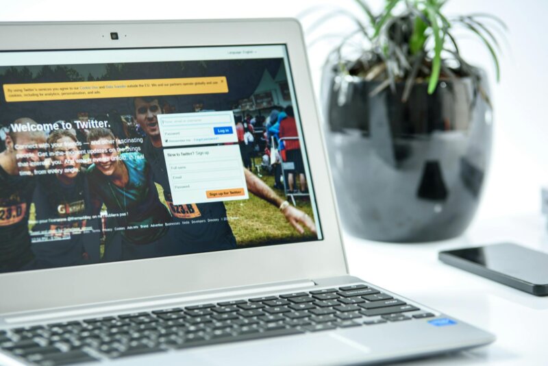 Close-up of a laptop displaying Twitter login page, accompanied by a smartphone and a potted plant in a bright workspace.