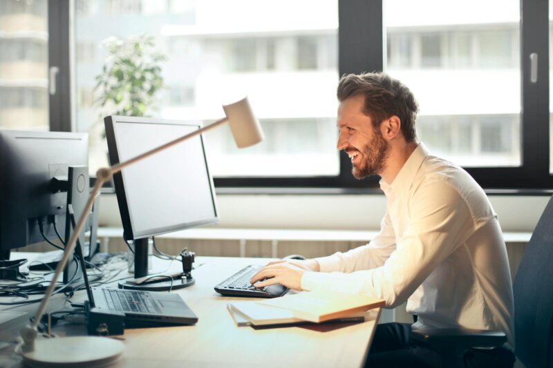 A man smiling while working at an office desk with a computer and natural daylight streaming in through large windows.